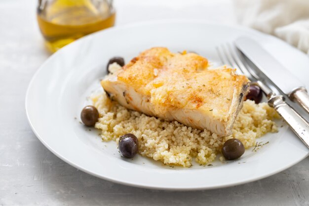 Fried cod fish with cous cous and olives on white plate