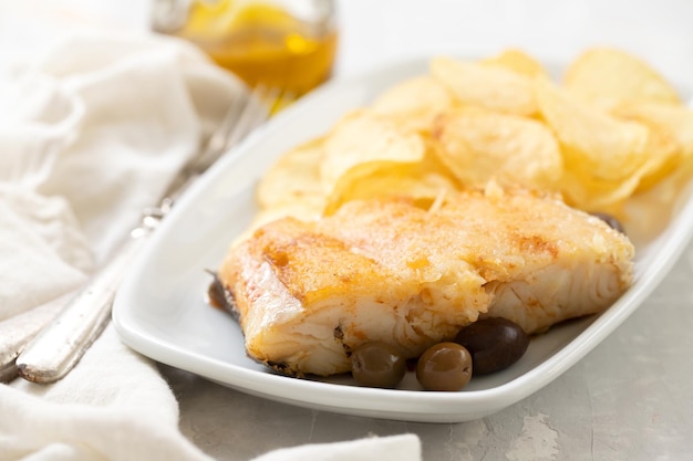 Fried cod fish with olives and potato chips on white dish on ceramic