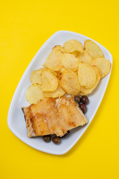 Foto baccalà fritto con olive e patatine su piatto bianco su fondo giallo