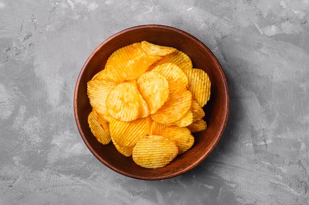 Fried corrugated golden potato chips in brown wooden bowl on concrete wall, top view