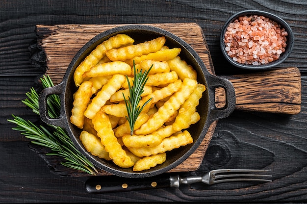 Fried Crinkle French fries potatoes in a pan. Black Wooden background. Top view.