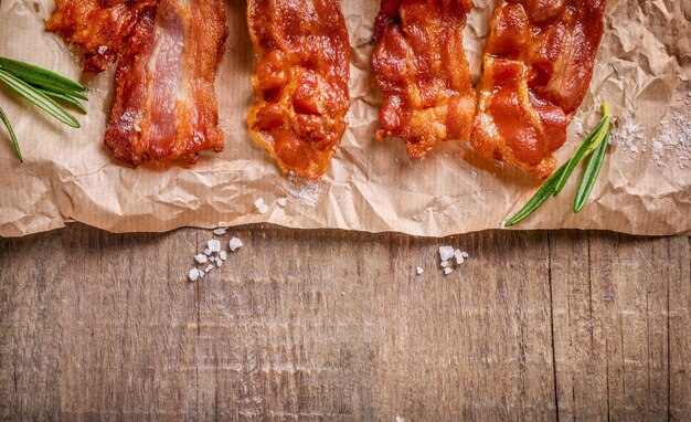 Fried crispy bacon slices, salt and rosemary sprigs on kraft paper on wooden