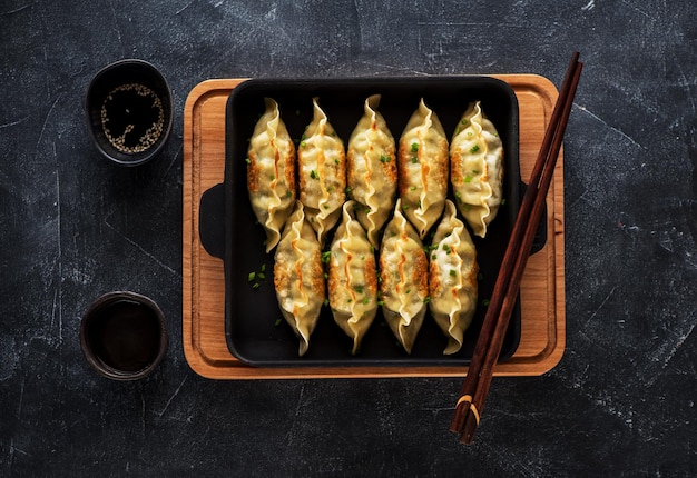 Жареные пельмени Gyoza на сковороде, соевый соус, палочки для еды на темном каменном фоне, вид сверху