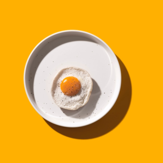 Fried egg on white plate on yellow with shadows.