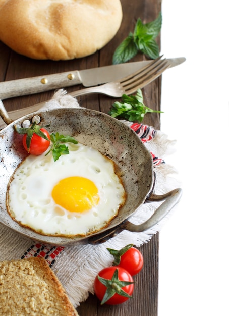 Fried egg with tomatoes, homemade bread and herbs on a old frying pan on wood close up