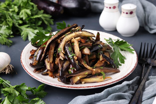 Fried eggplants with garlic and soy sauce, sliced in stripes, in a plate on a dark surface, closeup