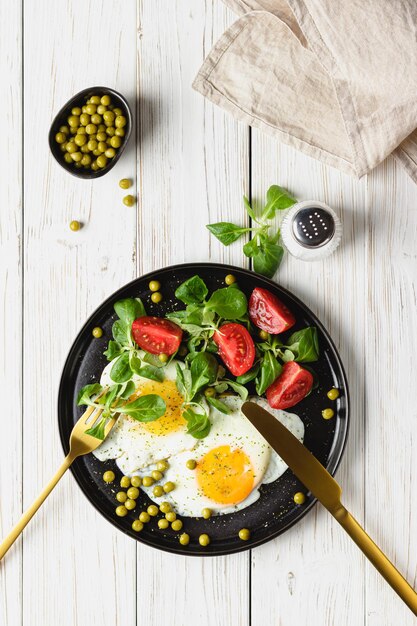 Fried eggs and salad with cherry tomatoes on black plate. Green peas in little bowl and salt in salt shaker. Homemade breakfast on white wooden table. Top view.