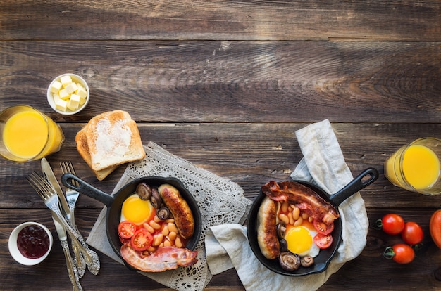 Fried eggs sausages bacon beans and mushrooms in iron skillet on rustic wooden background