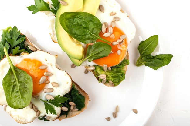 Fried eggs on a toasts bread with avocado, spinach and seeds on a white plate on the white background.
