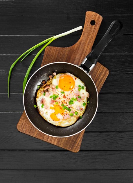 Fried eggs with ham and onion in pan on black wooden background Top view