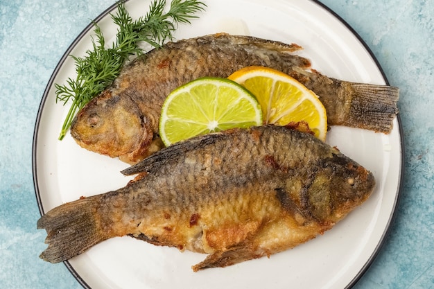 Fried fish Crucian on a plate with lemon and lime slices, top view. Ready meal. Blue background