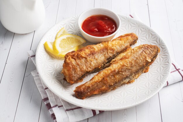 Photo fried fish hake pollock with tomato sauce and slices of fresh lemon. concept eating fatty fast food. white wooden table with copy space.