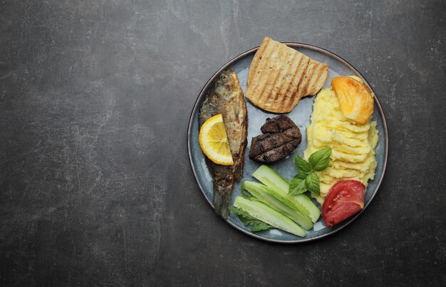 Fried fish, meat steak and vegetables on a plate. Concrete gray countertop