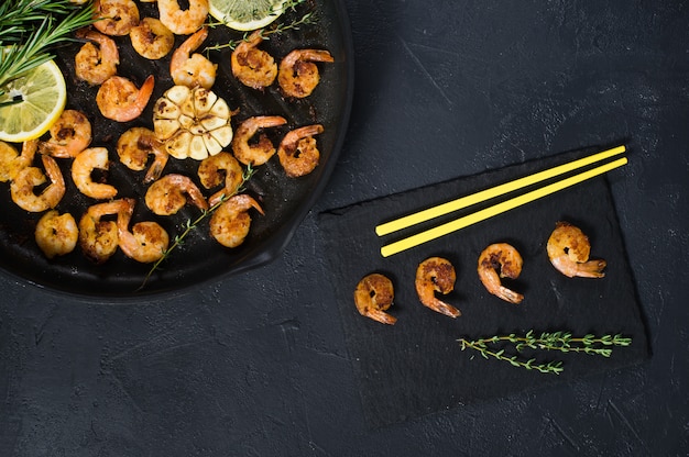 Fried king prawns in a frying pan on a black with yellow chopsticks.