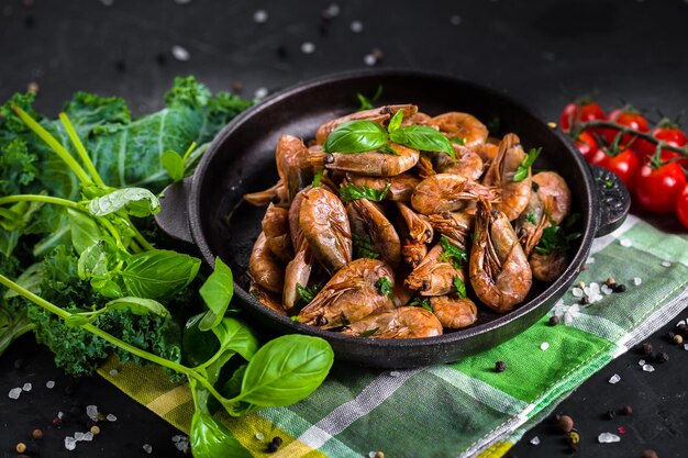 Fried large shrimps on a metal dish, a frying pan with herbs, vegetables with a cloth and a wooden