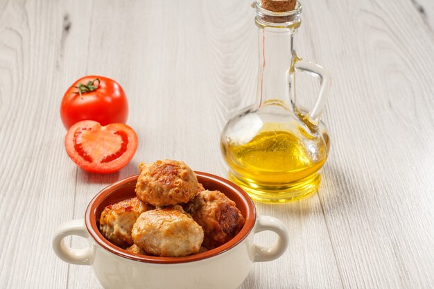 Fried meat cutlets in ceramic soup bowl, red tomatoes and glass bottle with sunflower oil on gray wooden boards.