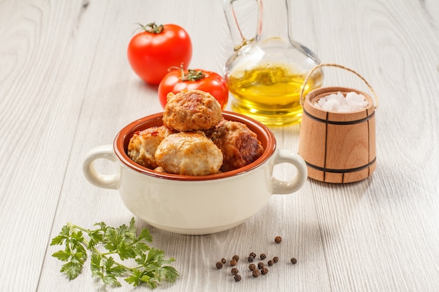 Foto cotolette di carne fritte in ciotola di zuppa di ceramica, pomodori rossi, bottiglia di vetro con olio di semi di girasole, cantina a botte di sale in legno e ramo di prezzemolo fresco su tavole di legno grigie.