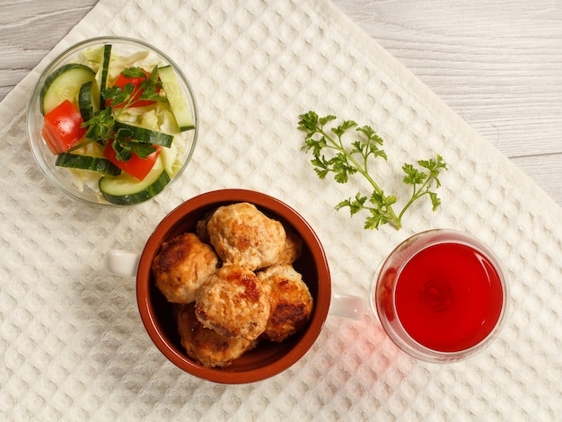 Fried meat cutlets in ceramic soup bowl, salad with tomatoes and cucumbers and glass of red wine on kitchen towel. Top view