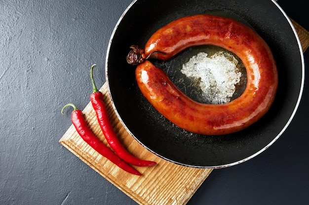 Fried meat sausage in a cast iron pan and hot chili pepper on a cutting wooden board on a dark stone table