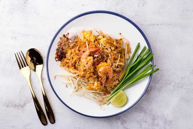 Fried noodle thai style with prawns in white dish and gold
spoon place on table. pad thai with vegetables (bean sprouts,
spring onions, lemon, tofu). popular street food of thailand. top
view.