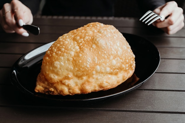 Fried pie with meat and onions on a black plate on woode table and hands holding fork and knife