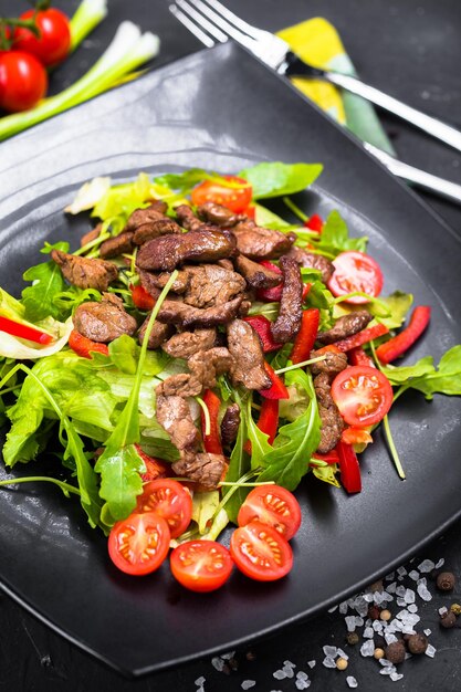 fried pieces of meat, on greens with cherry tomatoes, on a black plate, frying pan vegetables with