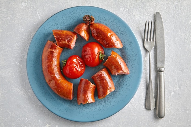 Fried pork sausage cut into pieces in a blue plate with tomatoes and cutlery on a light gray stone background