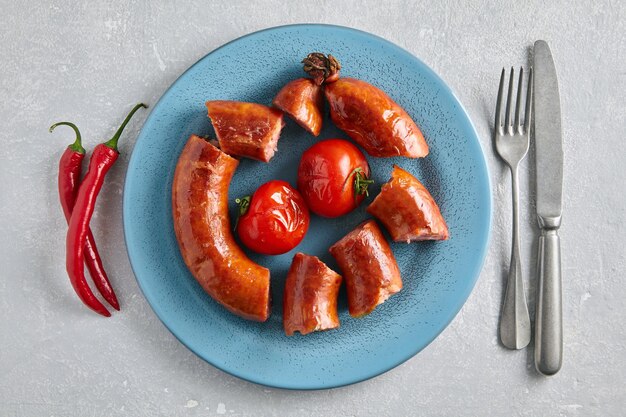 Fried pork sausage cut into portions and grilled tomatoes on a blue ceramic plate with cutlery and hot peppers on a gray concrete table Top view with copy space