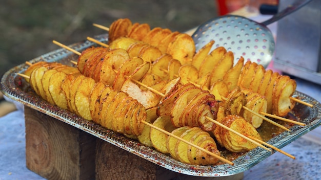 Fried potatoes in the form of spiral on wooden stick. Potatoes fried until golden color lies on metal tray during picnic. Potato chipsy take-away.