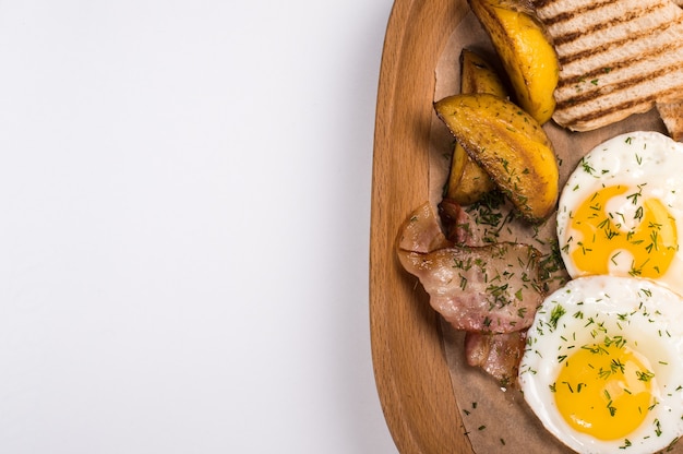 Fried potatoes with bacon, eggs and bread for breakfast, close up on wooden background. Top view with copy space