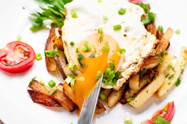 Fried potatoes with egg with liquid flowing yolk, herbs and tomatoes on a white plate.