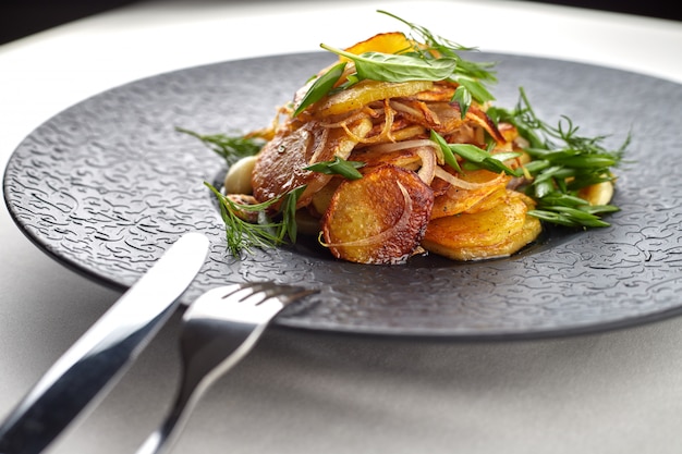 Fried potatoes with garlic and herbs on a black plate on a white surface