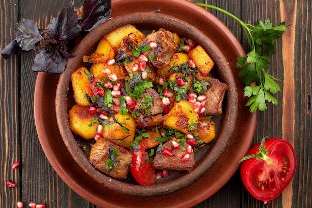 Fried potatoes with meat and pomegranate seeds, in a clay plate, on a wooden background with herbs and tomato