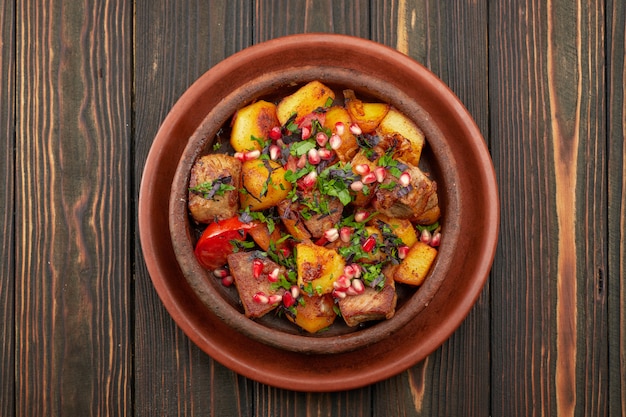 Fried potatoes with meat and pomegranate seeds, in a clay plate, on a wooden background