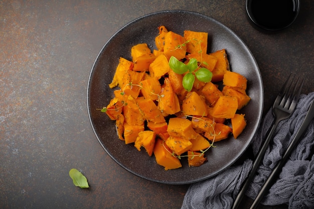 Fried pumpkin slices with herbs and pepper, olive oil in a black ceramic plate on dark concrete or stone