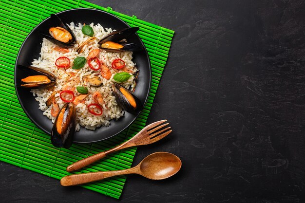 Жареный рис с мидиями из морепродуктов, креветками, базиликом в черной тарелке с деревянной ложкой и вилкой на зеленой бамбуковой циновке и каменном столе. Вид сверху.