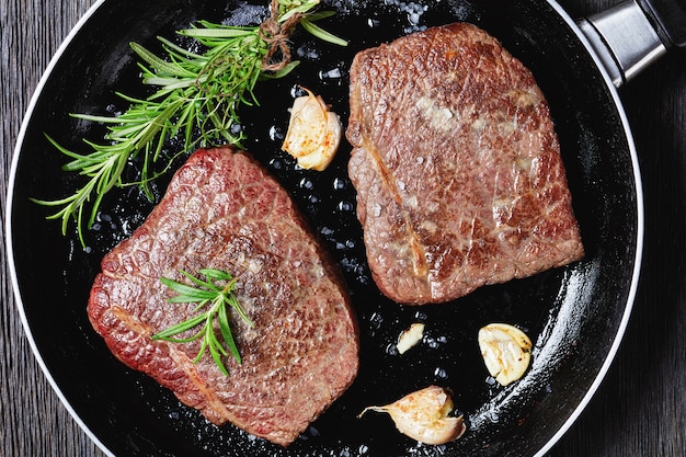Fried rump beef steaks with rosemary bouquet, garlic and salt on a skillet on a dark wooden table, horizontal view from above, flat lay, close-up