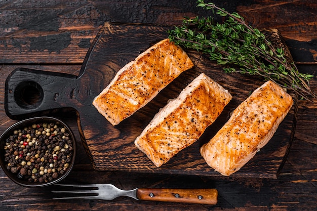 Photo fried salmon fillet steaks on a wooden board with thyme. dark wooden background. top view.