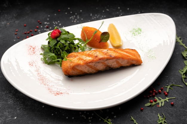 Fried salmon steak with herbs and lemon on a white plate on a dark background