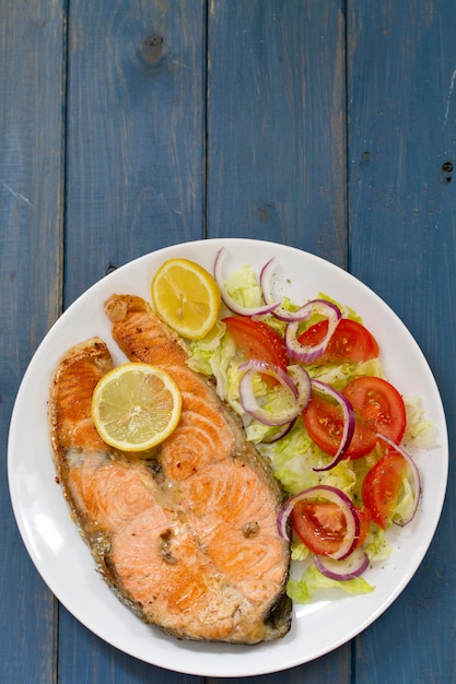 Fried salmon with vegetable salad and lemon on plate on blue wooden surface