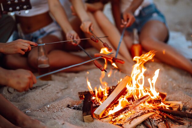 Fried sausages on bonfire Group of young friends sitting on beach and fry sausages Camping time
