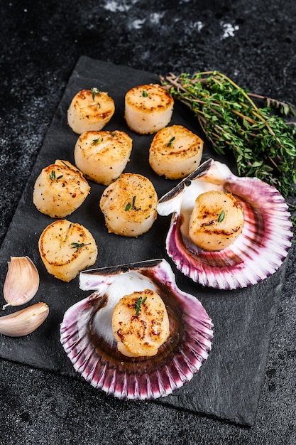 Fried seafood scallops meat with butter in a shells. Black background. Top view.