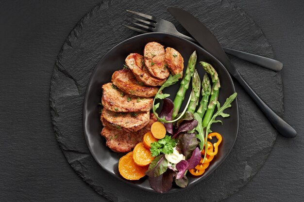 Fried slices of beef, sweet potato, asparagus and mixed green salad on black plate set for meal on dark slate