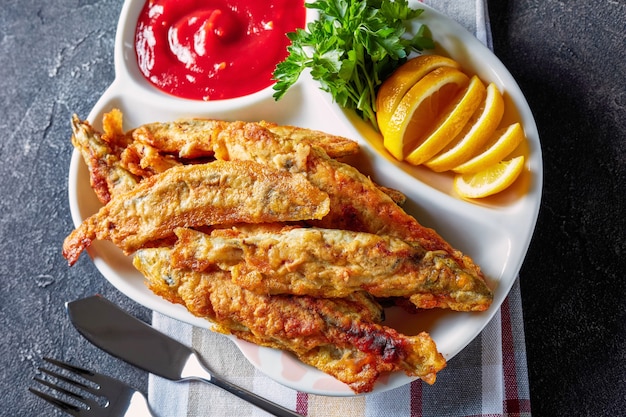 Fried small capelins with tomato sauce, sliced lemon on a plate on a concrete table with fork and knife, view from above, flatlay, close-up