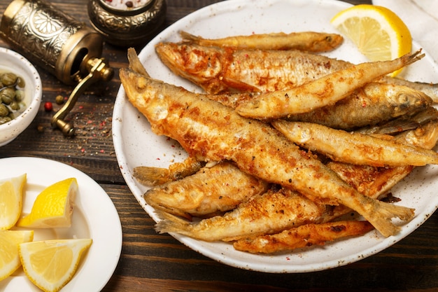 Fried smelt in a white plate, Small fish, Capers, lemon, pepper and salt on a wooden table, A delicious dinner in the rustic style