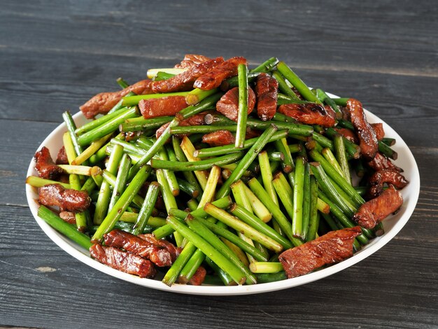 木製のテーブルの上の白いプレートに肉とニンニクの揚げ芽。中華料理。