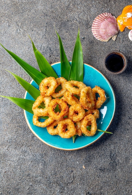 Photo fried squid rings on blue plate decorated with tropical leaves gray concrete background and sea decoration top view