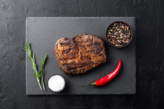 Fried steak on a black plate with spices and green sprigs of rosemary on a black table top view, flat lay of fried meat on a barbecue