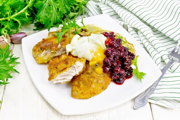 Fried turkey breast in breadcrumbs with cranberry sauce, boiled\
egg, baked parsnip and lettuce in a plate, napkin and fork on white\
wooden board background