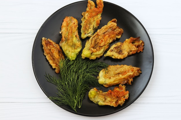 Fried zucchini flowers are located on a dark plate on a white surface, Top view